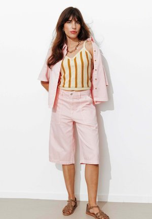 Femme debout contre un mur blanc, portant une chemise rose pâle, un short rose jusqu'aux genoux, un débardeur rayé et des sandales brunes.