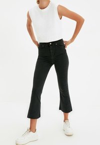 Svarta högmidjade jeans med utsvängda ben, en kort stickad vit ärmlös topp och vita sneakers. Har en diskret fransig fåll.