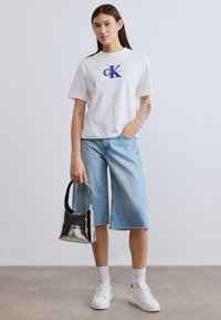 Λευκό βαμβακερό T-shirt με μπλε λογότυπο Calvin Klein, σε συνδυασμό με ανοιχτόχρωμες γαλάζιες denim culottes και λαμπερή μαύρη τσάντα· λευκά αθλητικά παπούτσια και κάλτσες.