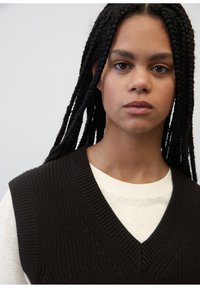 Svart strikket vest med V-halsdesign, som har teksturert stoff og ribbestrikkede detaljer, lagd over en kremfarget langermet skjorte.