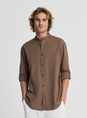 Młody mężczyzna o kręconych blond włosach, ubrany w brązową koszulę z kołnierzykiem typu mandarynka i podwiniętymi rękawami oraz białe spodnie, ręce w kieszeniach.