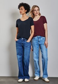 Due donne stanno fianco a fianco, indossando magliette a maniche corte; una di colore blu scuro, l'altra di bordeaux. Entrambe indossano jeans in denim a gamba dritta in diverse tonalità.