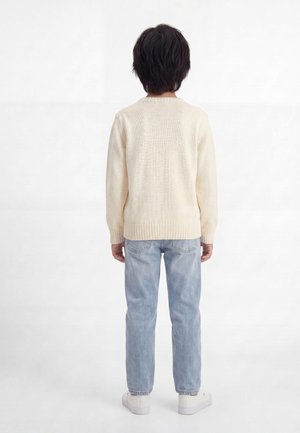 Enfant aux cheveux courts et foncés portant un pull en maille crème, un jean bleu clair et des baskets blanches, debout de dos sur fond blanc.