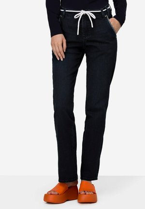 Jeans de mezclilla oscura con cintura elástica blanca y cordón, diseño ajustado, costuras en contraste y acentos azules, combinados con zapatos de plataforma naranjas.