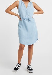 Světle modré šaty z chambray s výstřihem do V, knoflíkovou légou a stahovacími šňůrkami v pase. Bezrukávový design s zaobleným lemem. V kombinaci s černými teniskami.