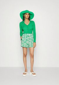 Cardigan verde texturizado com botões, combinado com calções verdes com estampa floral. A modelo usa um chapéu verde de aba larga e sandálias brancas de plataforma.