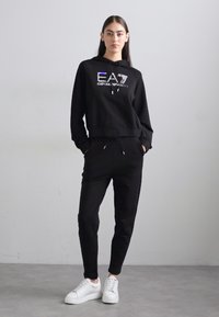 Juodas džemperis ir džinsai, su atspindinčiu EA7 logotipu. Lygus audinys su suspaustomis rankovėmis ir reguliuojamais laižais. Suderinta su baltais sportbačiais.