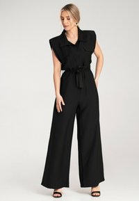 Czarny kombinezon z kołnierzykiem, rękawami typu cap, zapięciem na guziki, pasem w talii i szerokimi nogawkami. Gładki materiał bez widocznych wzorów.