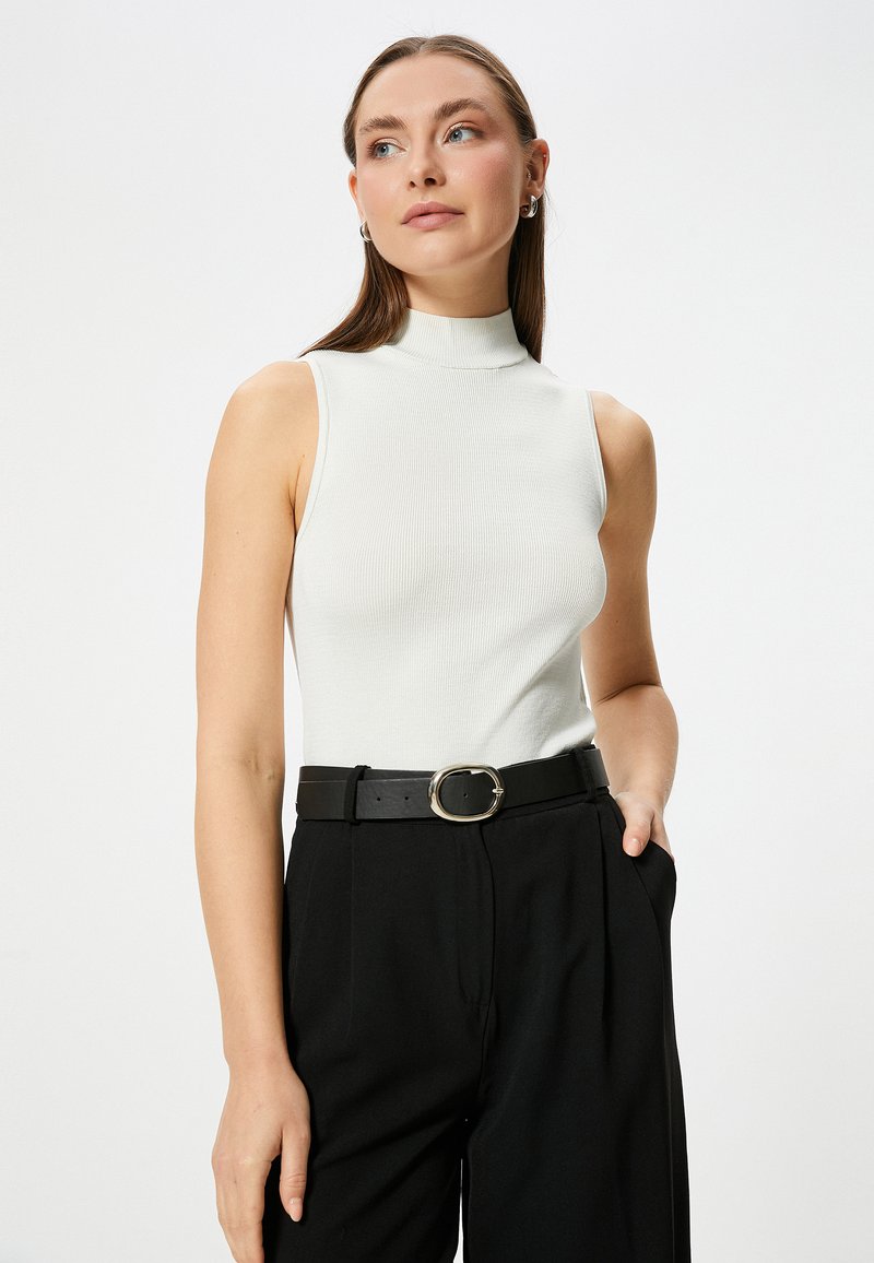 Witte ribbelige mouwloze top met een hoge halslijn, gecombineerd met hoge zwarte broeken en een zwarte riem met een ronde zilveren gesp.