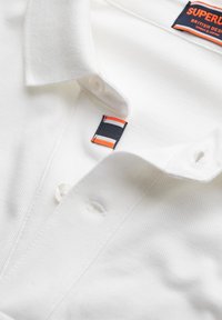 Weißes Polo-Shirt mit strukturiertem Gewebe, ausgestattet mit einer Zwei-Knopf-Leiste, einem Markenetikett und einem dekorativen gestreiften Akzent am Kragen.
