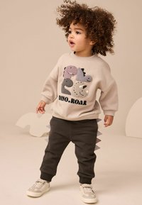 Felpa beige con motivi di dinosauro e testo "DINO.ROAR", abbinata a pantaloni scuri con dettagli a punta e scarpe da ginnastica bianche.