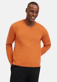 Maglione a V in un brillante colore arancione, realizzato in materiale testurizzato con polsini e orlo a coste, abbinato a pantaloni scuri.