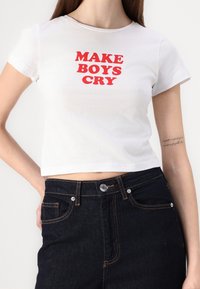 Camiseta corta de algodón blanca con texto rojo "HAZ LLORAR A LOS CHICOS" en el frente, combinada con unos jeans oscuros de cintura alta.