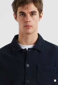 Junger Mann mit kurzen braunen Haaren, der ein dunkles, strukturiertes Hemd über einem weißen T-Shirt trägt und mit dem Blick zur Kamera gegen einen schlichten Hintergrund steht.