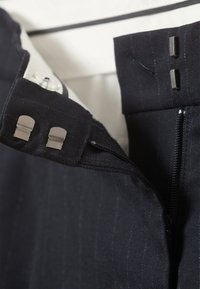 Czarne spodnie materiałowe z prążkowanym wzorem, z zamkiem błyskawicznym, metalowymi akcentami oraz zapięciem na guzik w pasie.