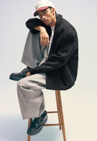 Black oversized blazer, white t-shirt, grey wide-leg trousers, black shoes with metal hardware, and a beige and burgundy cap, seated on a stool.