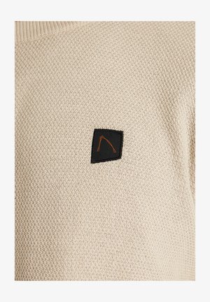 Bézs kötött pulóver textúrált mintával, amelyen a mellrészen egy fekete téglalap alakú folt található narancssárga körvonal-tervvel.