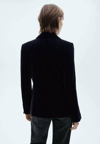 Sort fløjl blazer med klapkrave, to-knaps lukning og lange ærmer; glat tekstur og tætsiddende design.