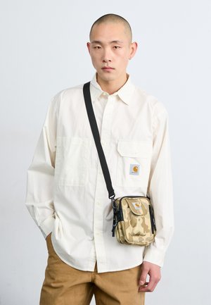 Jeune homme portant une chemise blanche ample et un pantalon beige avec un sac bandoulière camouflage beige sur un fond clair uni.