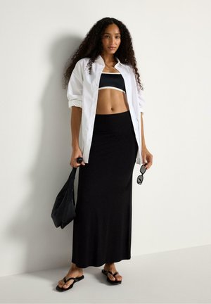 Kvinde med langt krøllet hår iført en hvid skjorte over en sort crop top, sort maxinederdel, sorte sandaler, holder en taske og solbriller.