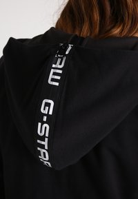 Felpa nera con cappuccio caratterizzata da una cerniera visibile sul retro e dal logo G-Star bianco a contrasto. Tessuto morbido, vestibilità comoda e superficie liscia.