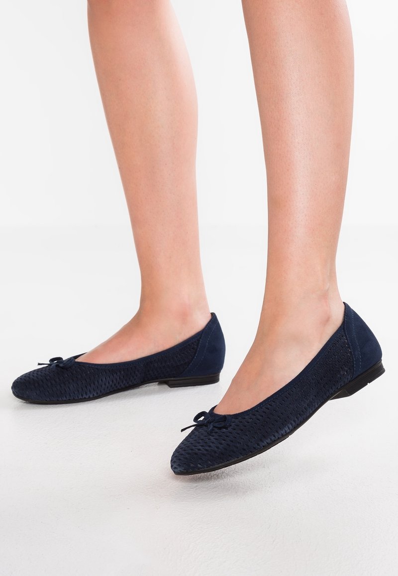 Pieds portant des ballerines bleu marine texturées avec un petit détail de nœud sur un sol blanc lisse.
