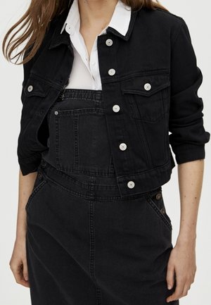 Mujer con una chaqueta vaquera negra recortada sobre una camisa blanca con cuello y un peto vaquero negro con bolsillo frontal y detalles de botones.