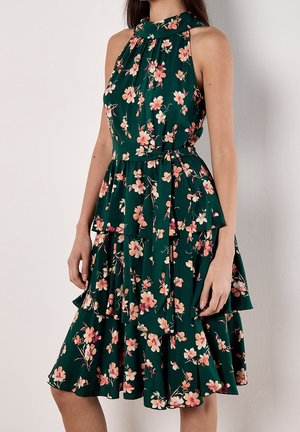 Vestido verde oscuro sin mangas con escote alto, falda en capas y estampado floral rosa, llevado por una persona de pie sobre un fondo liso.