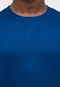 Κοντινό πλάνο ενός ατόμου που φοράει μπλε μπλούζα με στρογγυλή λαιμόκοψη και μικρό, μαύρο λογότυπο Calvin Klein στο κέντρο του στήθους.