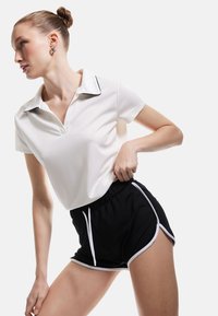 Polo shirt bianco a maniche corte con colletto nero, abbinato a pantaloni corti neri con rifiniture bianche e coulisse. Tessuto liscio e design aderente.