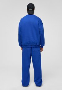 Blauwe sweater en bijpassende broek van zachte stof. Comfortabele pasvorm met geribbelde mouwen, een achterzak op de broek en een zwarte pet.