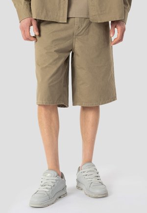 Mann trägt beige knielange Shorts und hellgraue Turnschuhe, steht vor einem schlichten hellgrauen Hintergrund.