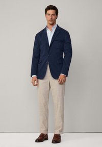 Blazer de algodón azul marino con solapas de muesca, un bolsillo en el pecho y dos bolsillos laterales, combinado con pantalones beige y mocasines marrones.