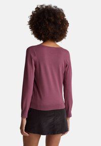 Person mit lockigem Haar, trägt ein langärmeliges mauvefarbenes Oberteil und einen schwarzen Minirock, steht mit dem Rücken zum Betrachter.