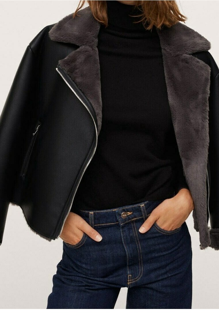 Veste noire en simili cuir avec col en fausse fourrure grise, fermeture éclair asymétrique, et manches larges, portée par-dessus un col roulé noir et un jean bleu.