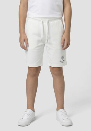 Enfant portant un short blanc à cordon avec poches latérales et petit logo sur la jambe gauche, associé à des baskets blanches sur fond uni.