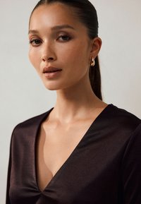 Crni top od satena s dubokim V-izrezom, glatke teksture i suptilnog sjaja. Zlatne naušnice u obliku obruča upotpunjuju izgled.