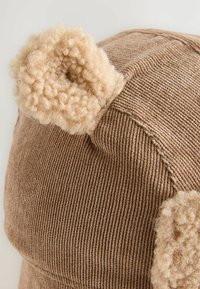 Brun ribbad corduroy-hatt med texturerade tanfärgade fuskpälsöron. Rund form med horisontellt ribbat mönster. Noterbara mjuka plyschdetaljer på öronen.