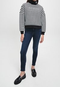 Sudadera a rayas en blanco y negro con cuello alto simulado, puños negros, combinada con jeans oscuros ajustados y mocasines negros brillantes.