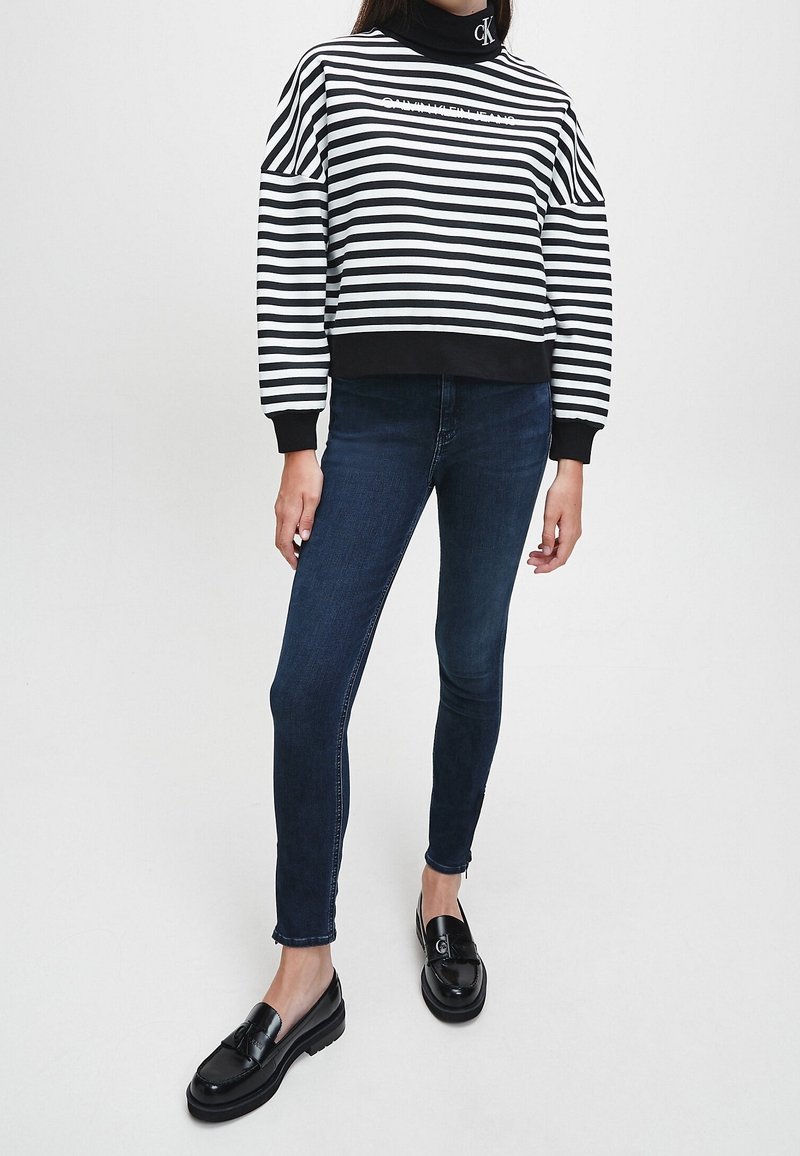 Sudadera a rayas en blanco y negro con cuello alto simulado, puños negros, combinada con jeans oscuros ajustados y mocasines negros brillantes.