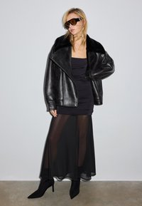 Czarna, oversize kurtka ze skóry z kołnierzem z sztucznego futra, dopasowana czarna sukienka, prześwitująca czarna spódnica i czarne szpilki z ostrymi noskami. Brązowe okulary przeciwsłoneczne.