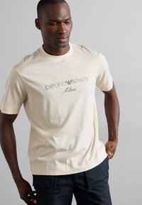Cremefarbendes T-Shirt mit kurzen Ärmeln aus Baumwolle, mit grauem bedrucktem Logo. Gerader Schnitt mit klassischem Rundhalsausschnitt.