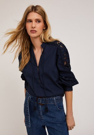 Marineblauwe blouse met een mandarinakraag, decoratieve kantdetails op de mouwen en een knoopsluiting aan de voorkant. Gecombineerd met high-waisted denim broeken.