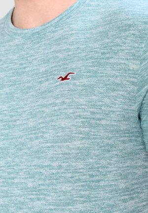 Hellblaues strukturiertes T-Shirt mit Rundhalsausschnitt und kurzen Ärmeln, mit kleinem rotem Vogel-Logo auf der linken Brust.