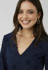 Robe marine avec un col en V, ornée de broderies florales, de petits boutons et de manches longues. Des boucles d'oreilles ovales dorées complètent le look.