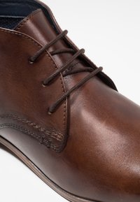 Scarpa in pelle marrone con una superficie liscia, dotata di lacci marrone scuro, punta arrotondata e cuciture dettagliate lungo il bordo.