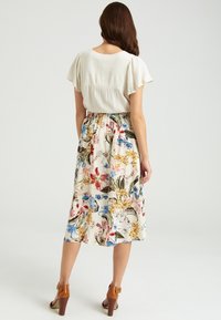 Bloemen midi-jurk met een crèmekleurige rok met kleurrijke botanische prints, korte pofmouwen en een geraffineerde taille. Bruine haksandalen.