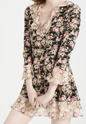 Frau trägt ein schwarzes Blumenkleid mit pinken und gelben Blumen, gerafftem Kragen sowie transparenten, gerafften Manschetten und Saum.