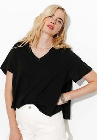 T-shirt noir à manches courtes en V, fabriqué en tissu doux, avec une coupe décontractée et des détails de couture discrets, associé à un pantalon blanc.