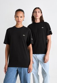 Két modell fekete Lacoste pólót és világoskék farmert visel, egy sima fehér háttér előtt állnak.